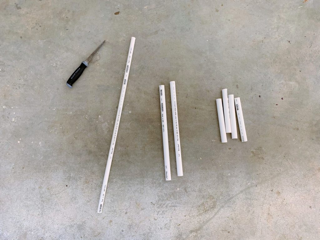 Seven sections of PVC pipe of varying sizes and diameters and a handsaw laying on the ground